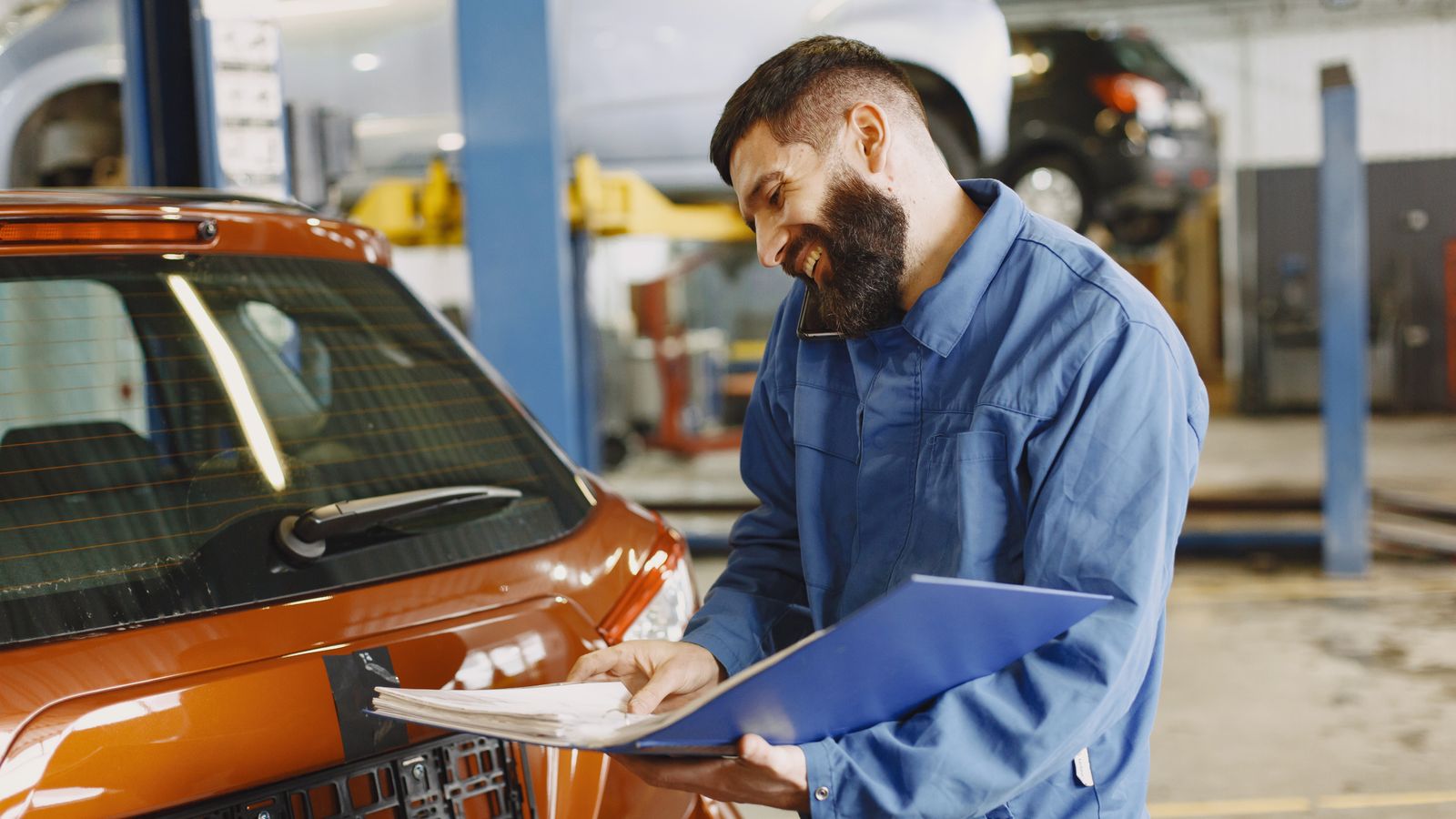 Mecánico sonriente con mono azul junto a un coche naranja en el taller